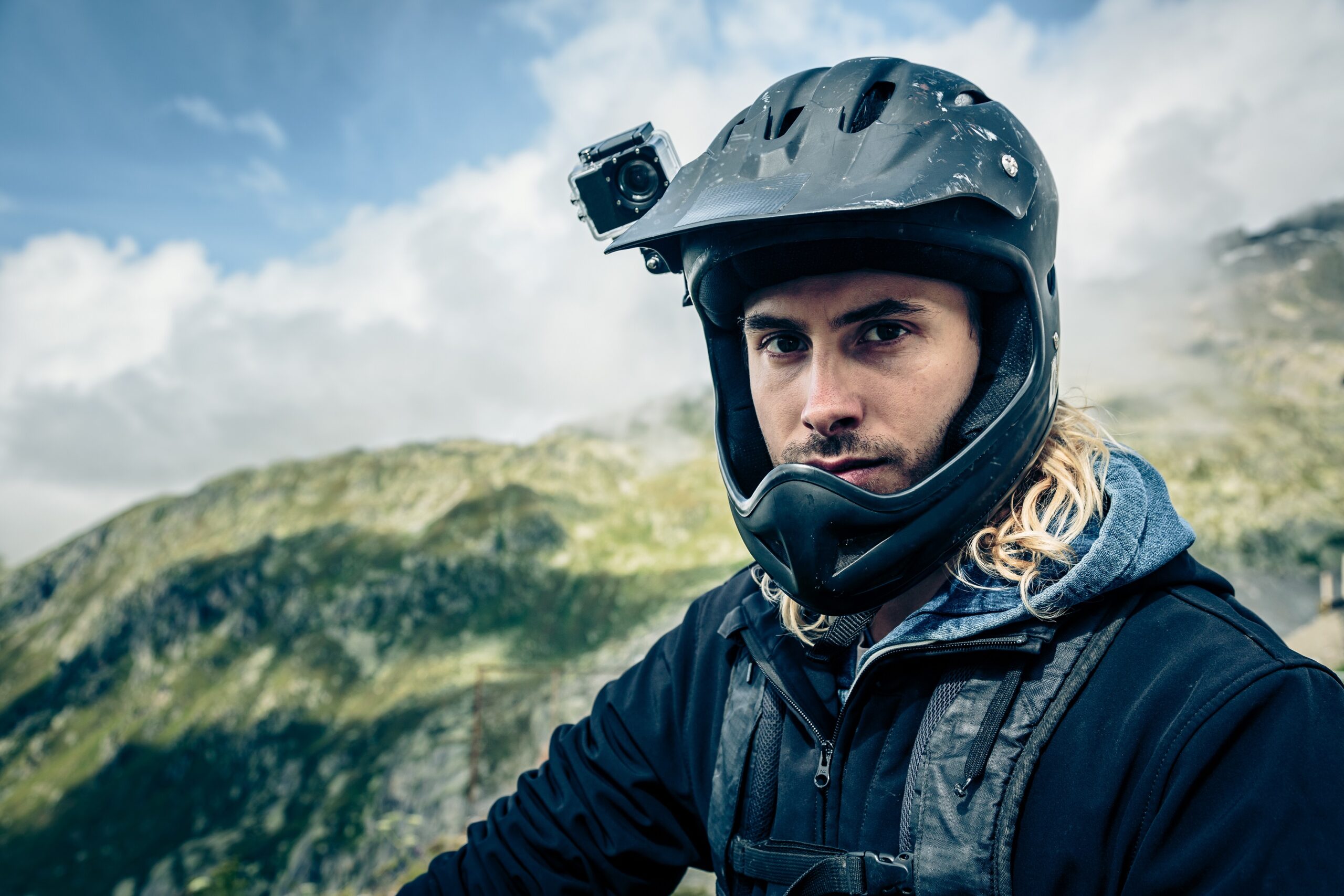 黒いヘルメットにカメラを取り付けた外国人の男性が晴れた空と山を背景にしてこちらを見ている