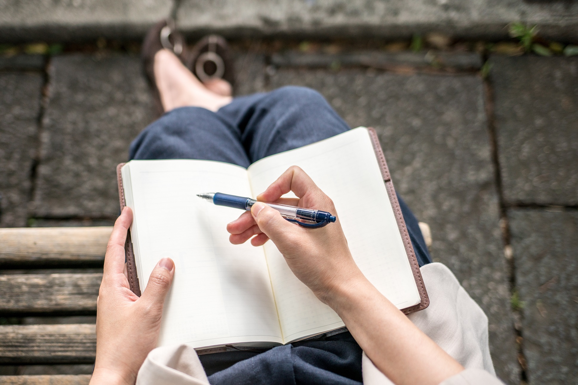 パンツとパンプスを履いた人が座って足の上にノートを広げなにか書こうとしている