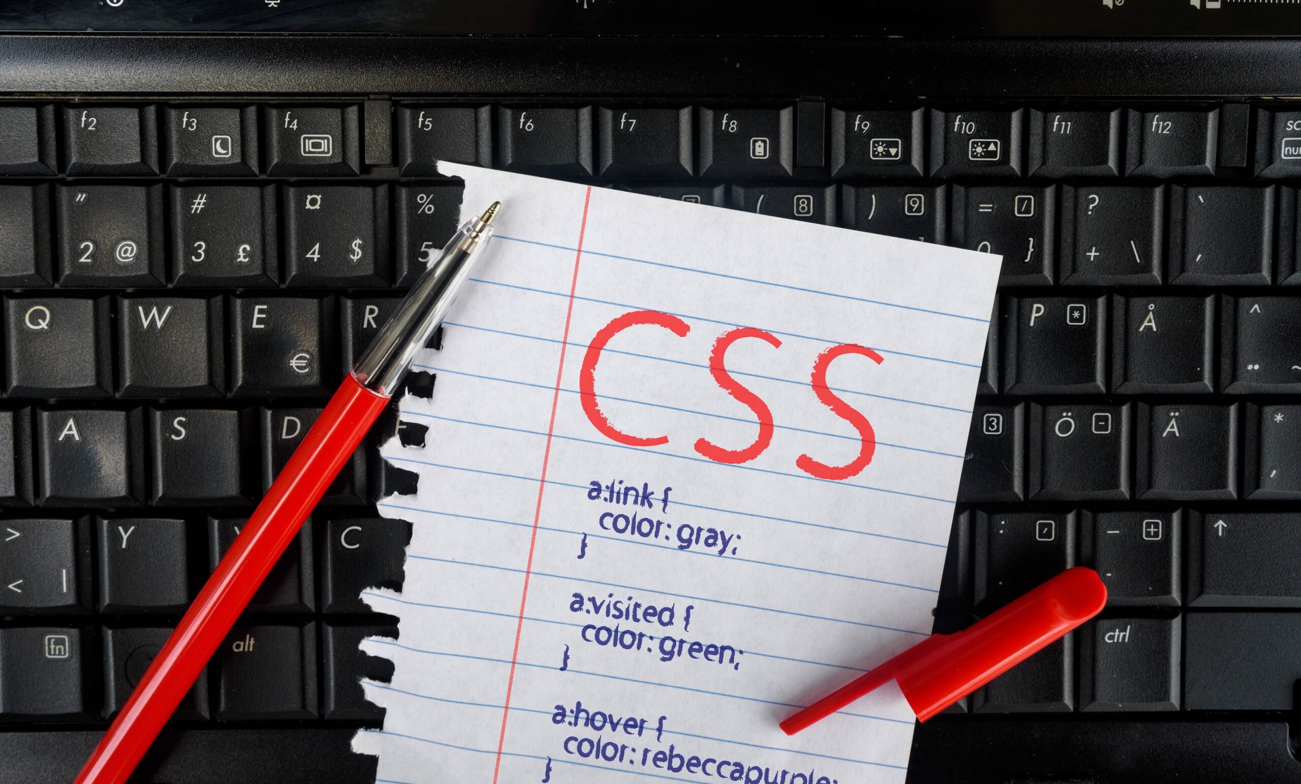 黒いキーボードの上に赤文字でCSSと書かれたメモ帳とペンが置かれている